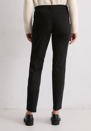 Person trägt eng anliegende schwarze Hose, beigen Pullover und schwarze Schuhe und steht mit dem Rücken zur Kamera vor einer schlichten hellen Wand.