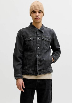 Jeune homme portant un bonnet en tricot beige, une veste en denim noire avec boutons, une chemise beige et un pantalon noir, debout devant un fond clair uni.