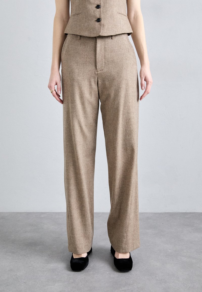Pantalones de cuadros marrones de tiro alto con un diseño de pierna recta, que cuentan con un cierre frontal y bolsillos laterales, combinados con zapatos negros de tipo slip-on.