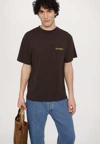 Braunes Baumwoll-T-Shirt mit einem gelben "NoProblemo"-Grafik auf der linken Brust, kombiniert mit blauen Jeans und einer braunen Tragetasche.