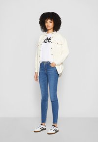 Giacca bianca con due tasche sul petto sopra una maglietta graficata bianca, abbinata a jeans skinny blu e sneakers bianche con strisce nere.