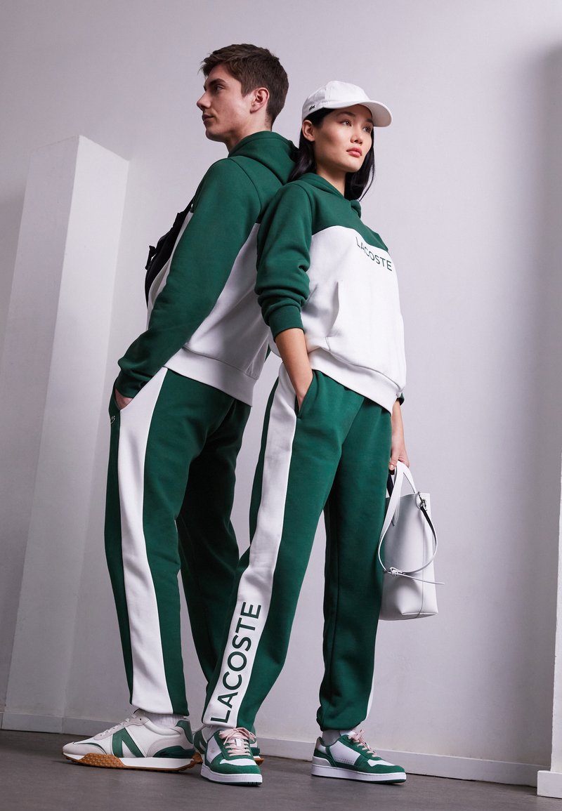 Felpa e pantaloni della tuta Lacoste verdi e bianchi, con strisce laterali e logo. Accessori includono un cappellino bianco e una borsa. Sneakers nei toni del verde.