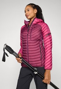 Chaqueta de plumas magenta con cuello alto, costuras en zigzag, bolsillos laterales y logo en la manga. La modelo sostiene bastones de senderismo negros.