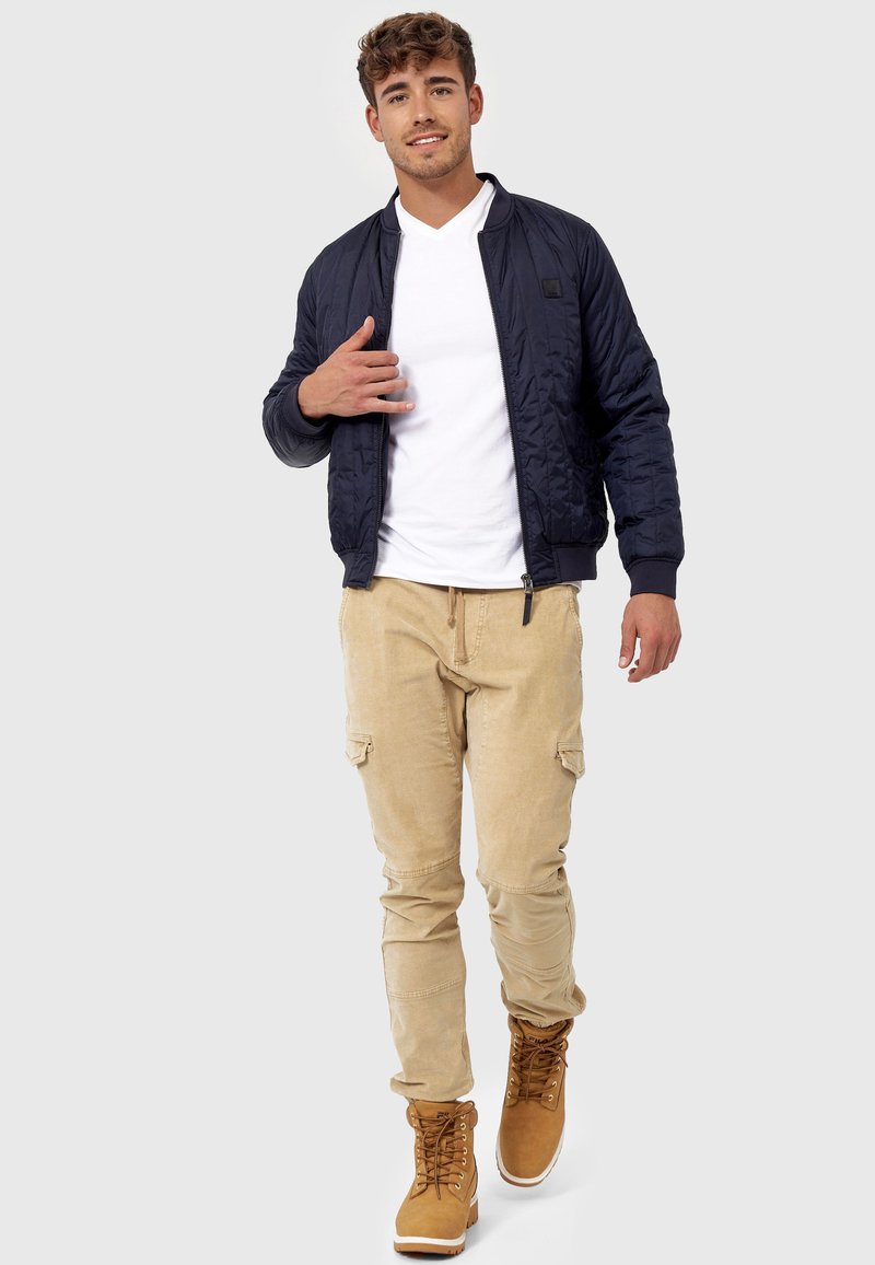 Mørkeblå quiltet bomberjakke, hvid V-hals t-shirt, beige cargobukser og beige snørebåndsstøvler. Minimalistisk design med lomme detaljer.