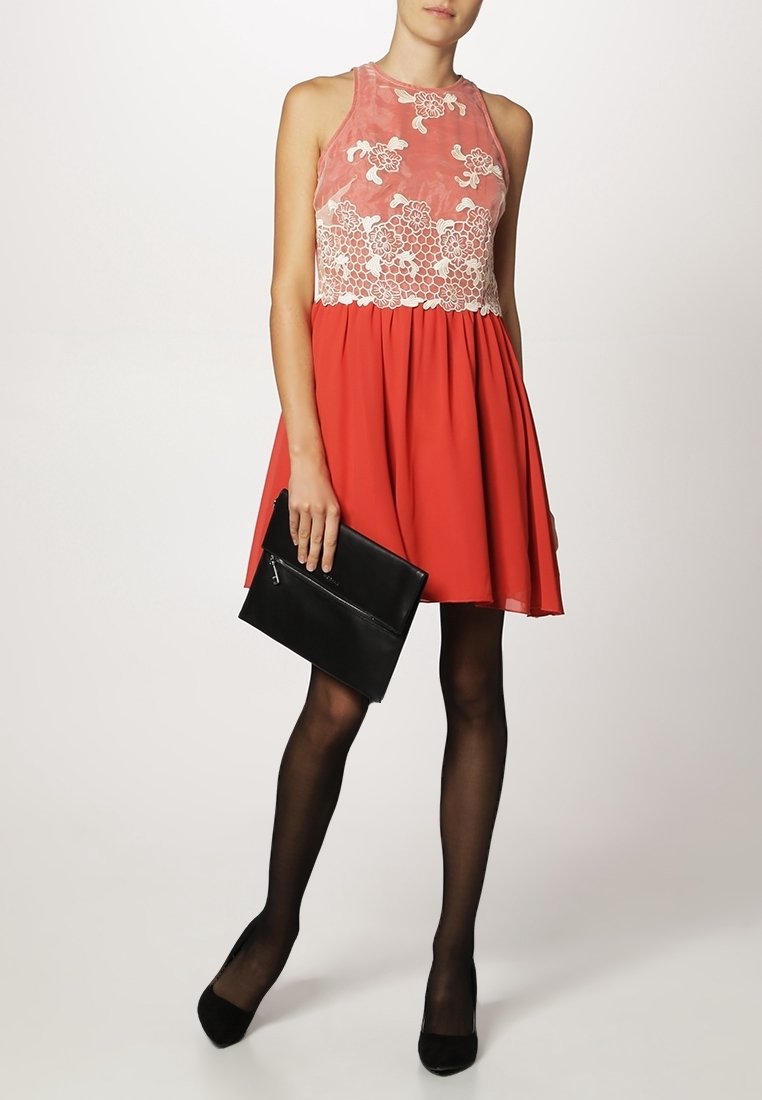 Červené šaty s přiléhavým bezramenním korzetem s květinovými krajkovými detaily, rozšířenou sukní a hladkým povrchem, doplněné černou klapkou.