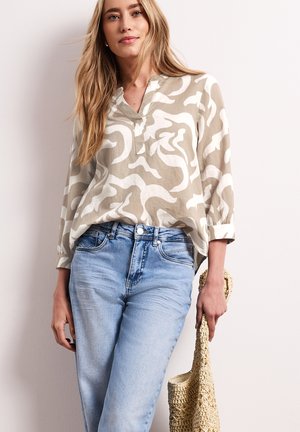 Młoda kobieta w beżowej i białej bluzce we wzory oraz jasnoniebieskich dżinsach, trzymająca plecioną beżową torebkę przy boku.