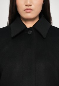 Manteau en laine noir avec un grand col et une fermeture à un bouton. Texture douce avec une finition lisse et uniforme et un minimum de détails.