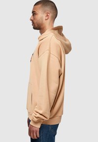 Beige bomullshuvtröja med avslappnad passform, känguruficka och ribbade muddar, sedd från sidan mot en neutral bakgrund.