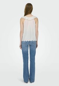 Femme debout de dos portant un chemisier blanc ajouré sans manches avec un col à volants et un jean bleu droit sur fond uni.