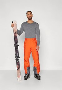 Costum de schi format dintr-un tricou gri cu mâneci lungi și pantaloni impermeabili portocalii strălucitori cu accente gri. Ține schiuri cu legături negre.