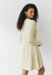 Robe pull en crème, avec un motif en cable, des manches longues et une finition évasée. Fabriquée en tissu doux avec des détails texturés.