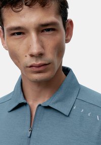 Világoskék, zipzáras, galléros ing, texturált anyagból, finom mintázattal a vállán és minimalista dizájnelemekkel.