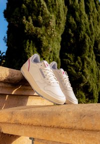 Kremowe sneakersy z perforowanym czubkiem, szarym zamszowym wykończeniem, różowymi akcentami i białymi sznurówkami, spoczywające na kamiennej półce.