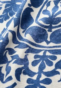 Tissu blanc à texture tissée présentant un motif floral et de feuilles bleues en répétition, montré avec des plis et des ombres.