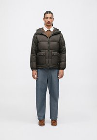 Svart boblejakke med glidelås foran, teksturert stoff og to brystlommer; paret med vide blå jeans og brune sko.