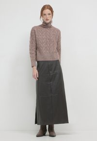 Frau in einem mauvefarbenen, kabelgestrickten Rollkragensweater, langer schwarzer Lederrock mit Seitenschlitz und braunen Stiefeletten, die vor einem weißen Hintergrund steht.