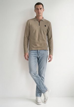 Mand stående op ad en hvid væg iført beige langærmet polo, lyseblå jeans og hvide sneakers, med benene krydsede og hænderne afslappet langs siderne.