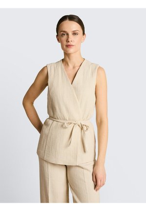 Frau mit braunem zurückgebundenem Haar, die ein ärmelloses beiges Wickeltop mit gebundenem Gürtel und passende weite Hosen trägt.