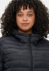 Melns pūkains mētelis ar augstu apkakli, ar quilted tekstūru un priekšējo rāvējslēdzēju. Audums izskatās gluds un viegls.