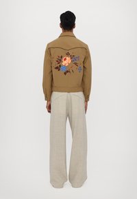 Hnědá bunda s květinovou výšivkou na zadní straně; doplněná světlými, volnými lněnými kalhotami. Texturovaná tkanina a dobře padnoucí design.