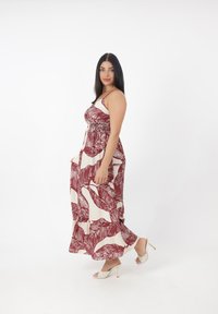 Robe maxi avec un imprimé de feuilles rouge et beige. Présente des fines bretelles, un corsage smocké et une silhouette fluide. Portée avec des talons à motifs.