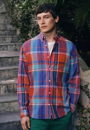 Young man wearing colorful plaid shirt and green pants stands near stone stairs with greenery in the background, hands relaxed by sides.