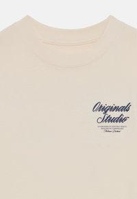 Light beige cotton t-shirt with a round neckline and short sleeves. Features a navy blue text logo on the left chest. Smooth texture.