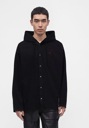 Jeune homme aux cheveux courts et foncés porte un sweat à capuche noir boutonné avec un petit logo rouge, debout devant un fond clair uni.