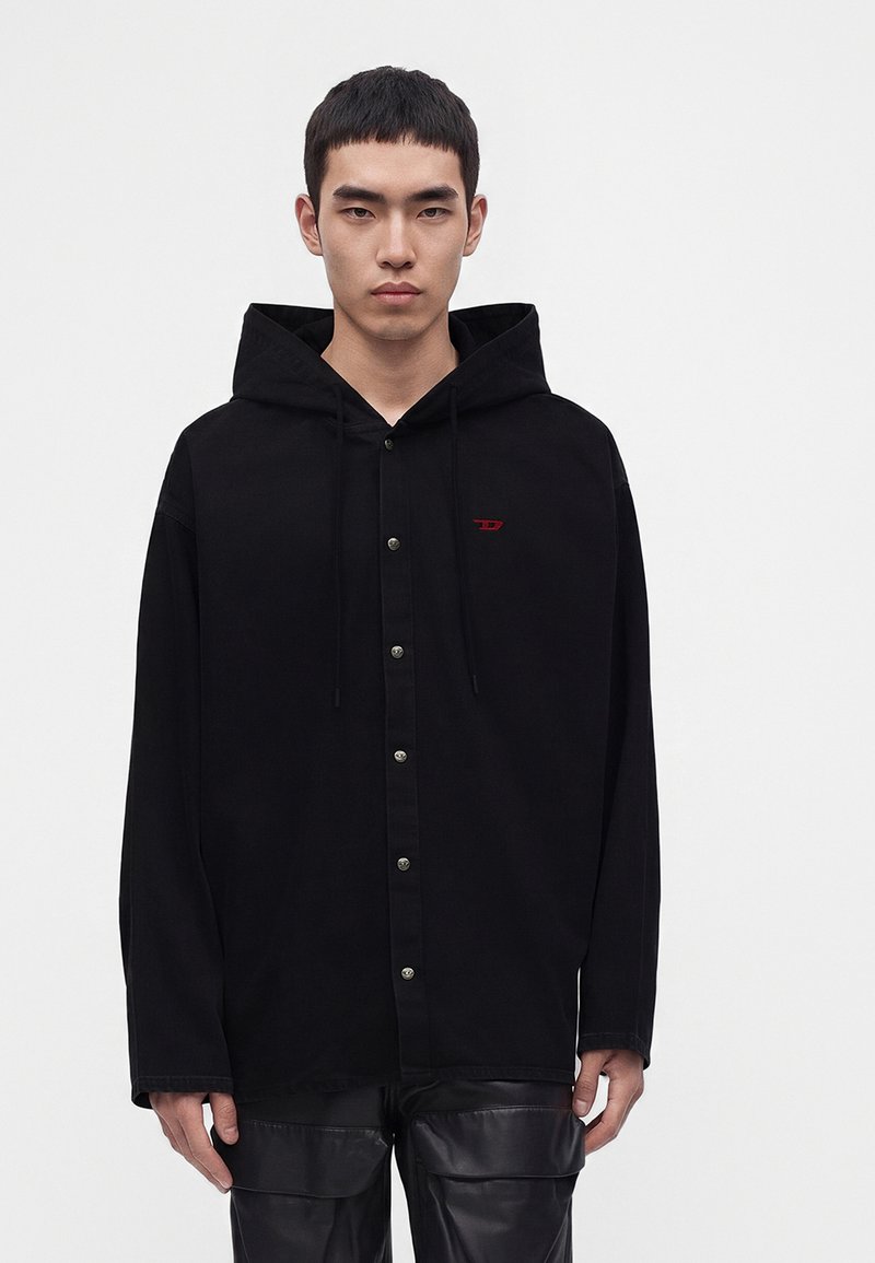 Jeune homme aux cheveux courts et foncés porte un sweat à capuche noir boutonné avec un petit logo rouge, debout devant un fond clair uni.