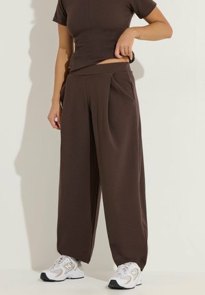 Osoba nosząca brązowe spodnie z szerokimi nogawkami i pasującą brązową bluzkę z krótkim rękawem, regulująca talię, stojąca na białym tle w białych tenisówkach.