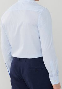 Chemise à carreaux bleu clair à manches longues, poignets boutonnés et texture lisse. Portée avec un pantalon bleu marine avec une poche arrière.