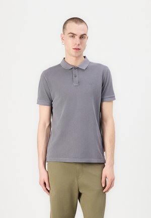 Graues Polohemd mit Kragen und einer Knopfleiste mit drei Knöpfen. Kurze Ärmel, weicher Stoff, besticktes Logo auf der Brust, getragen mit olivgrünen Hosen.