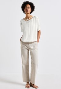 Weiße, kurzärmelige Bluse mit strukturiertem Oberflächen und überkreuztem Ausschnitt, kombiniert mit hellbeigen, locker sitzenden Hosen und Sandalen.