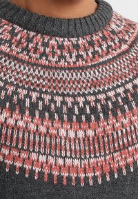 Pull gris avec un col rond, présentant un empiècement à motif en rouge et blanc. La texture est tricotée, mettant en avant un tissu dense.