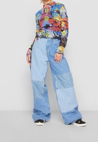 Buntes Langarmoberteil mit Smiley-Gesicht und floralem Druck; blaue Jeans im weiten Schnitt mit Patchwork-Details; schwarz-weiße gemusterte Schuhe.