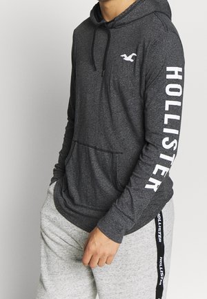 Personne portant un sweat à capuche Hollister gris foncé avec logo sur la poitrine et la manche, et un pantalon de survêtement gris clair avec une bande Hollister sur le côté.