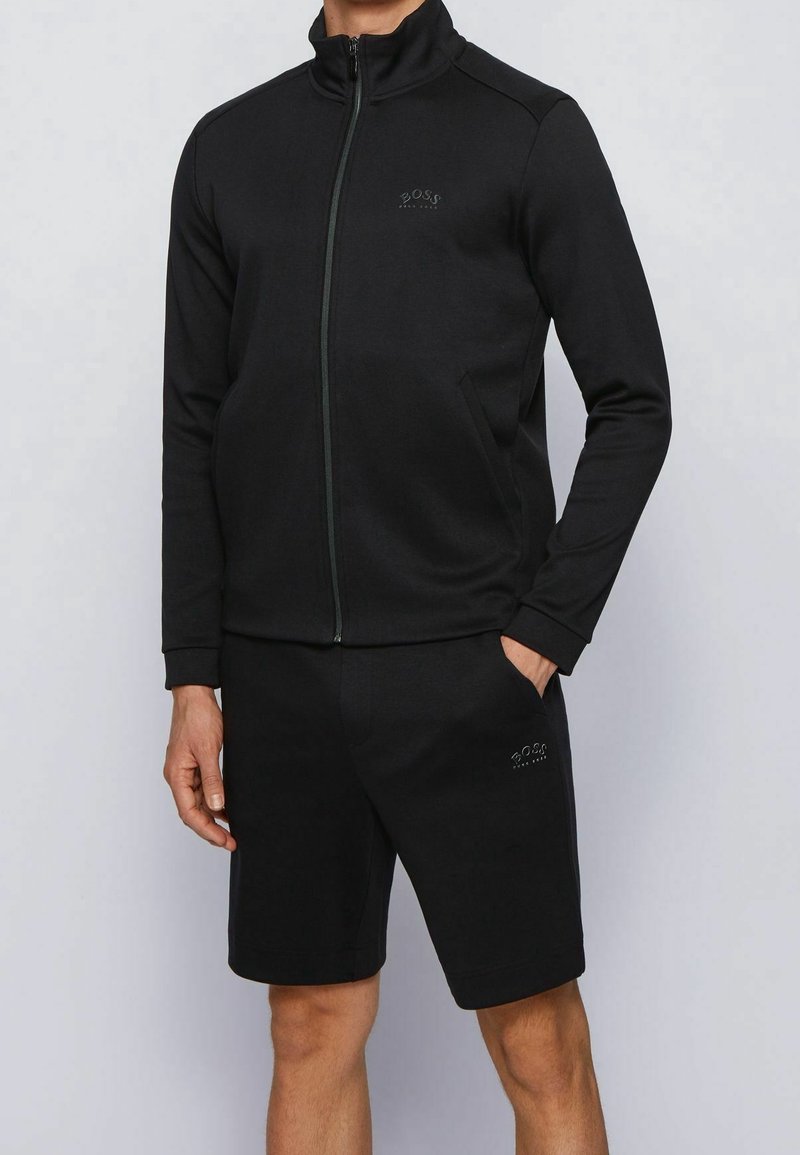 Tuta atletica nera composta da giacca con zip e colletto alto con logo, abbinata a pantaloni corti coordinati. Materiale morbido e design slim fit.