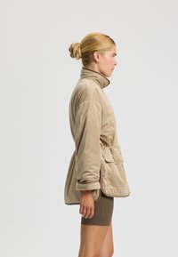 Chaqueta de pana beige con cuello alto, cintura ceñida con cinturón y bolsillos frontales; presenta un tejido texturizado y un corte relajado.