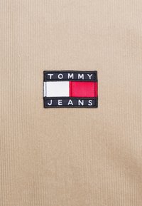 Tommy Jeans -logo, jossa on valkoinen, punainen ja musta väri beigestä corduroy-kankaasta.