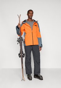 Άνδρας φορώντας πορτοκαλί και γκρι σακάκι σκι, μαύρο σορτς σκι και μποτάκια, κρατώντας ένα ζευγάρι σκι κατακόρυφα σε απλό άσπρο φόντο.