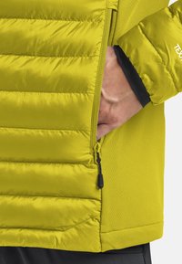 Mano in tasca con zip di giacca trapuntata gialla brillante, con pannelli laterali in morbido tessuto e polsino elastico su manica nera.