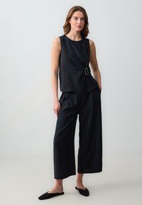 Haut sans manches noir avec un design drapé et un détail de boucle en or ; associé à un pantalon large noir et des chaussures noires à enfiler.