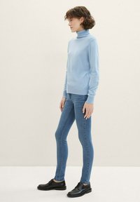 Suéter de cuello alto azul claro, de mangas largas, con puños y dobladillo acanalados. Combinado con jeans ajustados azul y zapatos de cordones negros.