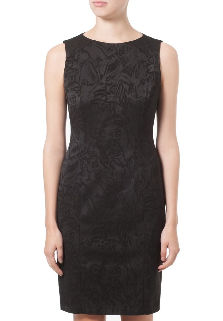 Femme portant une robe noire sans manches, longueur genou, avec des motifs subtils en texture et un col rond, posant devant un fond blanc.