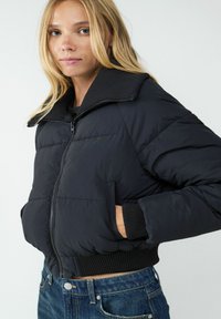 Czarna kurtka puchowa z wysokim kołnierzem, zapięciem na zamek i ściągaczami przy mankietach. Design zawiera pikowany wzór oraz boczne kieszenie.