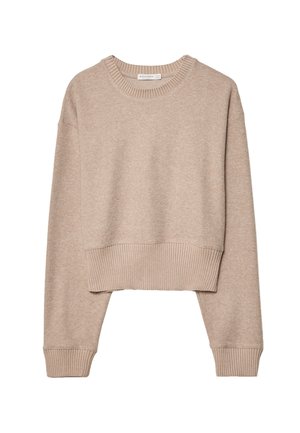 Personne portant un pull court beige et un pantalon noir taille haute à jambes larges, debout devant un fond rouge.