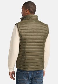 Olijfgroene geïsoleerde vest met verticale gestikte patroon, hoge kraag en logoaccent op de achterkant, gedragen over een lichtgekleurde trui.
