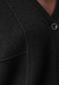 Svart kofta i texturerad tyg, med V-ringad design, knappstängning och synliga sömmar. Mjuk och varm känsla.