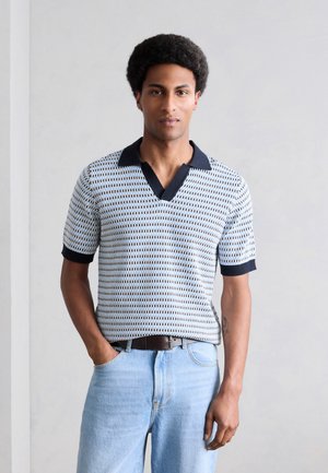 Junger Mann mit Afro-Frisur, der ein gemustertes kurzärmeliges Polohemd und hellblaue Jeans trägt und mit einer Hand in der Tasche steht.