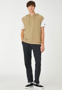 Beige mouwloze hoodie met trekkoorden over een wit shirt, gecombineerd met donkere gestructureerde broeken en zwarte sneakers. Eenvoudig ontwerp, casual pasvorm.